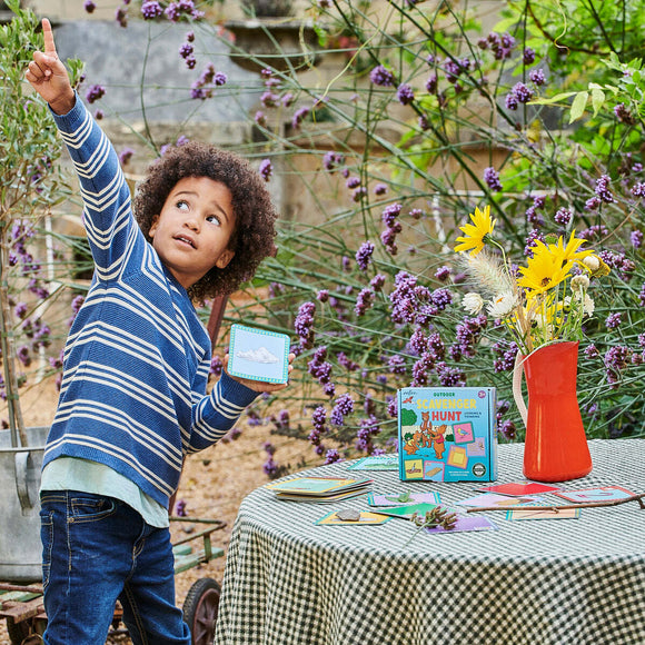 A young child with curly hair excitedly points to the sky while holding a card from the 'Scavenger Hunt Game - Outdoors' by Eeboo. The game box is visible on a checkered tablecloth alongside colorful cards and a bright red vase filled with flowers, set in a lush outdoor area surrounded by purple flowers.