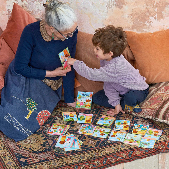 A grandmother and her grandson engage in a playful storytelling session using 'A Busy Day Create a Story Cards' by Eeboo. The cozy setting features colorful story cards spread out on a patterned rug, with the child excitedly pointing to a card as the grandmother smiles and listens. Decorative pillows and a textured wall add warmth to the scene.