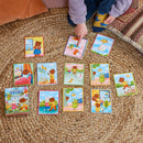 A child points to colorful story cards spread out on a textured mat. The cards depict various characters like bears and rabbits in vibrant, playful scenes. The game box is visible at the bottom left, titled 'Create a Story: A Busy Day' from Eeboo. The setting is cozy and inviting, perfect for imaginative play.