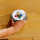 A child's hand reaches for a colorful, circular game device featuring four colored buttons (red, yellow, green, blue) and playful facial illustrations. The game is designed to engage kids in interactive play, enhancing cognitive skills through fun and learning. It rests on a wooden table, showcasing its vibrant design.