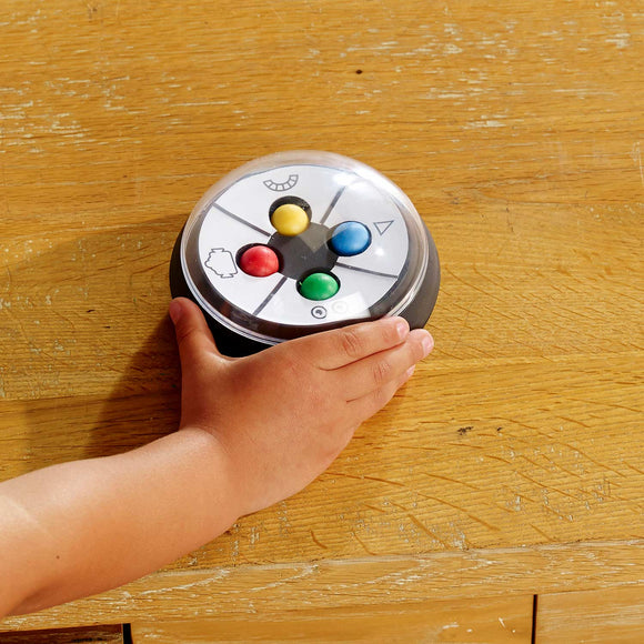 A child's hand reaches for a colorful, circular game device featuring four colored buttons (red, yellow, green, blue) and playful facial illustrations. The game is designed to engage kids in interactive play, enhancing cognitive skills through fun and learning. It rests on a wooden table, showcasing its vibrant design.