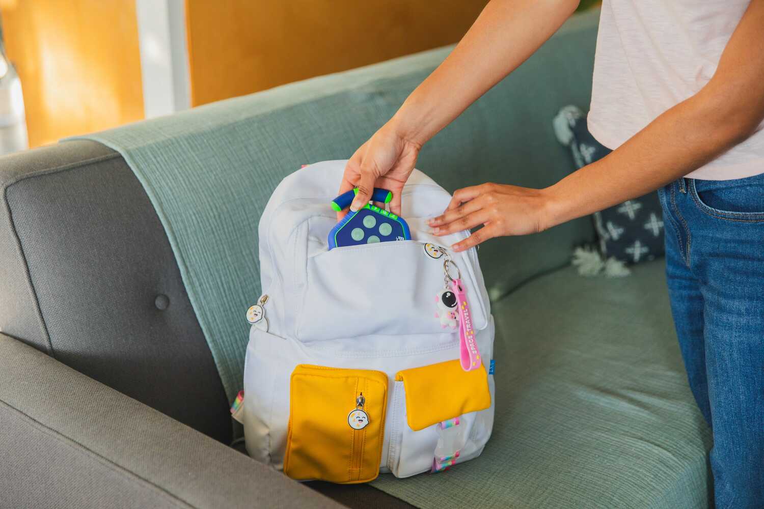 A person placing a BrainBolt Boost game into a white backpack with colorful pockets. The game features a blue and green design, while the backpack is positioned on a gray sofa, showcasing a casual and vibrant study environment.