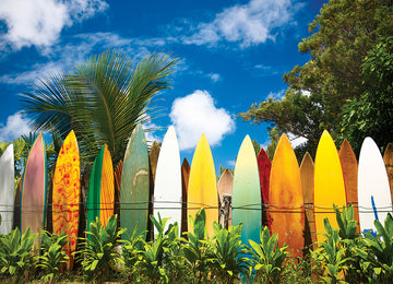 A vibrant scene of colorful surfboards standing against a wooden fence, surrounded by lush green plants. The backdrop features a bright blue sky with fluffy white clouds, evoking the spirit of surfing in Hawaii.