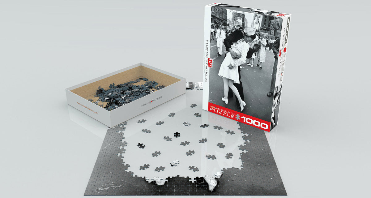 A 1000-piece jigsaw puzzle featuring the iconic V-J Day kiss in Times Square. The puzzle pieces are shown scattered around the partially assembled image, with the box displayed beside it, showcasing the historic moment captured in black and white.