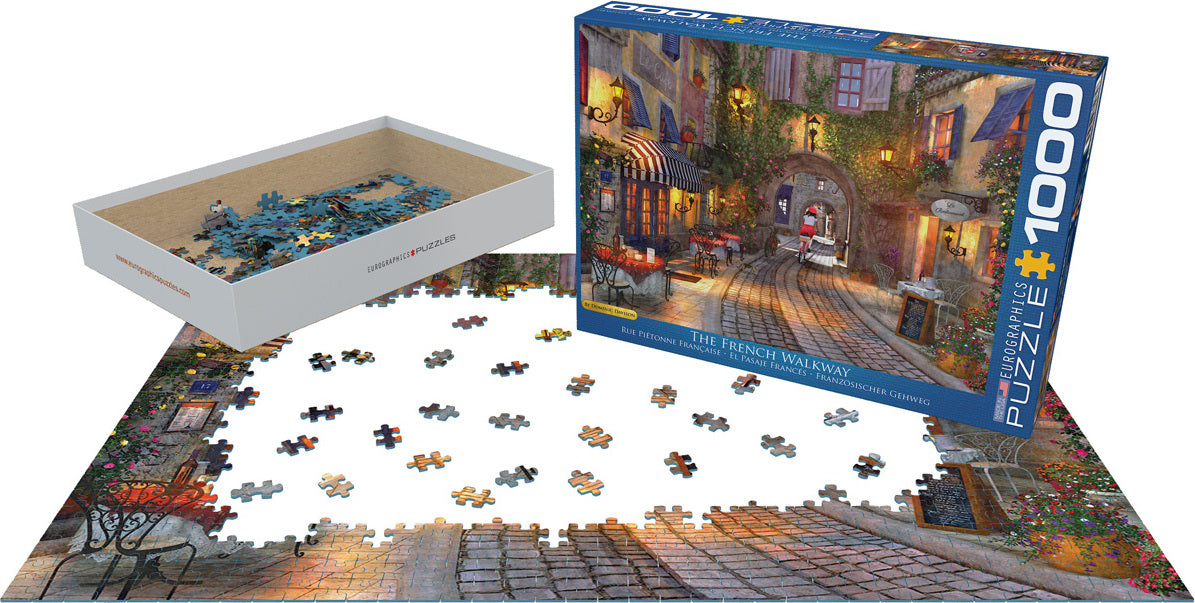 A jigsaw puzzle titled 'The French Walkway' by Dominic Davison, showing a picturesque street scene with vibrant colors and charming architecture. The box is displayed next to the puzzle with pieces scattered on a table. The puzzle features 1000 pieces and highlights outdoor cafes and cobblestone pathways.