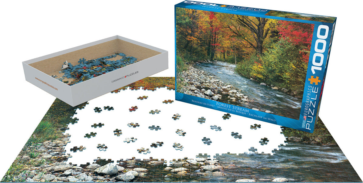 A 1000-piece jigsaw puzzle titled 'Forest Stream' by Eurographics, featuring a scenic view of a river surrounded by vibrant autumn trees. The pieces are scattered on a table, with the box displayed beside it, showcasing the completed image on the front.