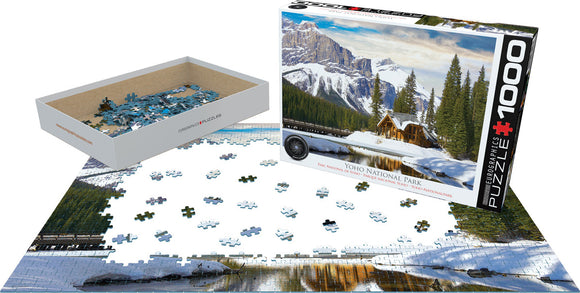 A jigsaw puzzle featuring a breathtaking winter scene of Yoho National Park in British Columbia. The image shows snow-capped mountains, evergreen trees, and a charming wooden cabin beside a serene lake. The puzzle consists of 1000 pieces and is packaged in a standard box with the image on display. Some puzzle pieces are scattered on a flat surface, demonstrating the progress of assembly.