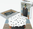 A jigsaw puzzle featuring René Magritte's iconic painting 'The Son of Man.' The image shows the assembled portion of the 1000-piece puzzle, depicting a man in a suit with an apple hovering in front of his face. The puzzle box is displayed beside it, showcasing the artwork on the cover and the array of scattered pieces inside the tray.
