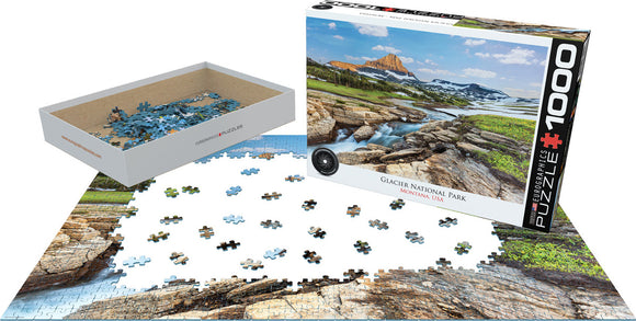 Eurographics 1000-piece jigsaw puzzle featuring a stunning landscape of Glacier National Park, Montana. The image showcases rocky formations, lush greenery, and scenic hills under a clear blue sky. The puzzle is displayed partially assembled, with scattered pieces visible in a white storage box.
