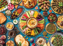 A vibrant and colorful Middle Eastern table spread featuring an array of traditional dishes. The centerpiece includes a bowl of hummus surrounded by fresh vegetables like carrots, cucumbers, and peppers. Various appetizers such as stuffed grape leaves, kibbeh, and pastries like sambousek are beautifully arranged, alongside olives, pomegranate seeds, and sweet desserts like baklava. The entire spread showcases a rich diversity of textures and colors on a blue tablecloth.