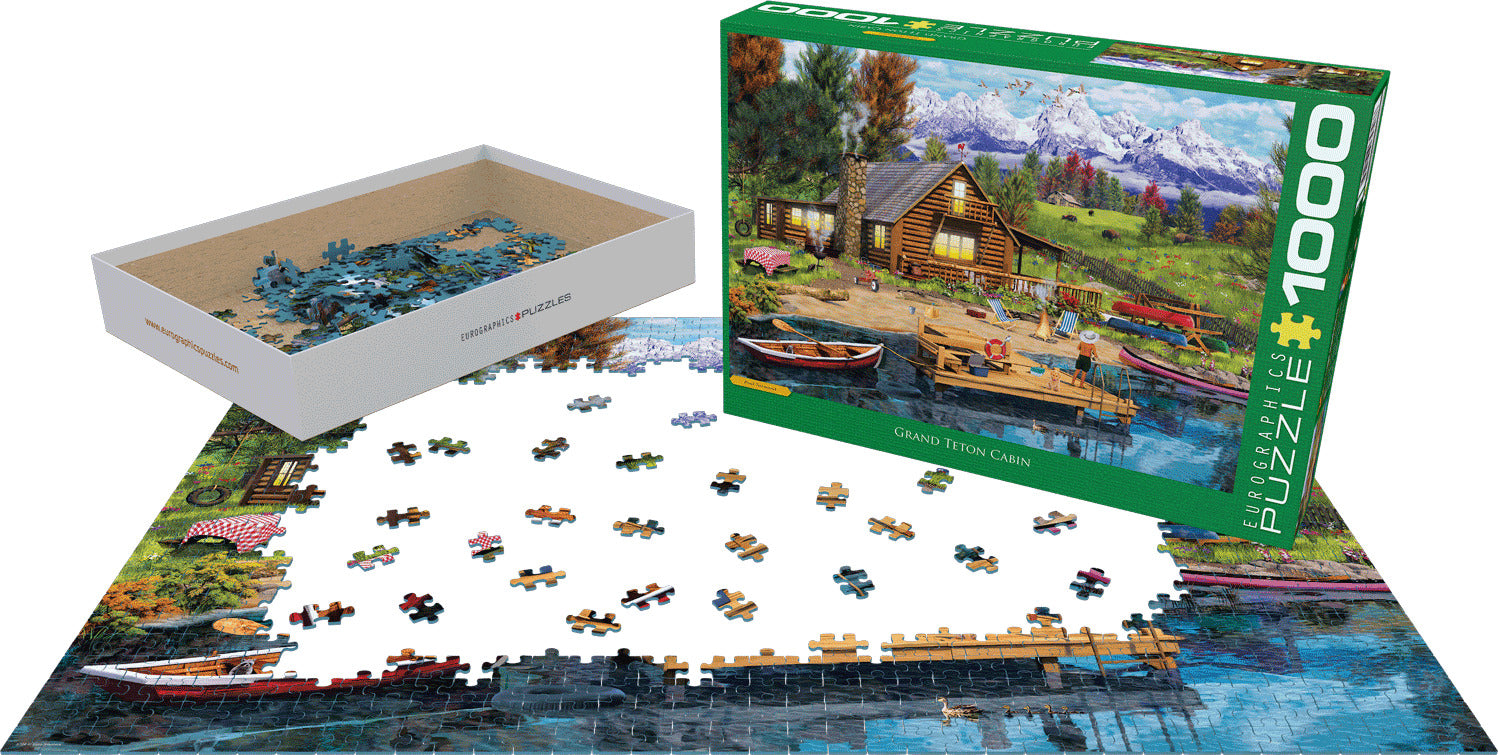 A 1000-piece jigsaw puzzle titled 'Mountain Cabin' by Paul Normand, featuring a serene mountain landscape with a cozy cabin by a lake. The puzzle pieces are displayed scattered around, with a partially completed image showing a boat and trees. The box contents include a beige tray with more puzzle pieces. EuroGraphics branding is visible on the box.