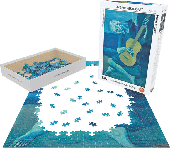 An intricate jigsaw puzzle of Pablo Picasso's 'Old Guitarist', showing several blue and gray pieces in a partially completed layout. The puzzle box sits nearby, displaying the artwork and details. This 1000-piece puzzle features the iconic image of an elderly man playing guitar, showcasing Picasso's signature style.
