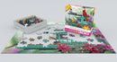 A vibrant jigsaw puzzle titled 'Bertie's Bird Seed Fly-In' from Eurographics. The image features colorful bird feeder surrounded by flowers, with puzzle pieces scattered around the partially completed scene and the puzzle box displayed nearby.
