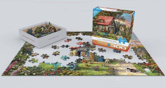 The Country Shed jigsaw puzzle by Dominic Da, displayed with its box. The scene features a charming country shed surrounded by vibrant flowers and greenery. A collection of colorful puzzle pieces is partially assembled on a flat surface, showcasing the beautiful artwork and intricate details. The box is positioned next to the puzzle, labeled with the title and '300 pieces'.