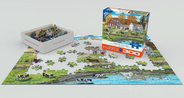 The 'Farm by the Lake' jigsaw puzzle by Bob Fair, produced by Eurographics. The image shows a partially completed puzzle featuring a serene rural scene with cows near a lake, alongside the puzzle box displaying the artwork. The puzzle pieces are arranged around the box, showcasing vibrant colors and intricate details.