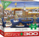 Packaging of the Seaside Holiday jigsaw puzzle by Carol Dyer, featuring a festive coastal scene with decorated boats, snow, and a bustling town. The puzzle consists of 300 XL pieces and is suitable for family fun. The box displays vibrant artwork, highlights the actual size of the pieces, and carries the Eurographics logo.