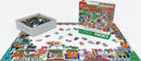 The Downtown Holiday Festival jigsaw puzzle by Eurographics features a colorful 500-piece design depicting a festive downtown scene filled with holiday decorations, shops, and people celebrating. The puzzle is partially completed on a table, with a box of loose pieces displayed beside it, showcasing large pieces that are easy to handle and assemble.