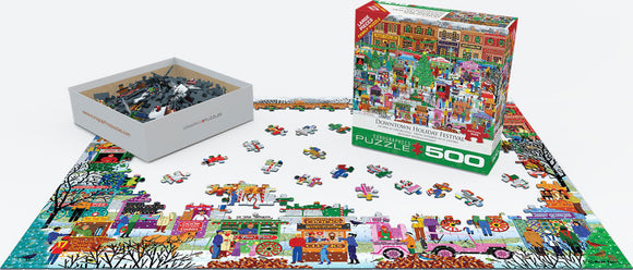 The Downtown Holiday Festival jigsaw puzzle by Eurographics features a colorful 500-piece design depicting a festive downtown scene filled with holiday decorations, shops, and people celebrating. The puzzle is partially completed on a table, with a box of loose pieces displayed beside it, showcasing large pieces that are easy to handle and assemble.