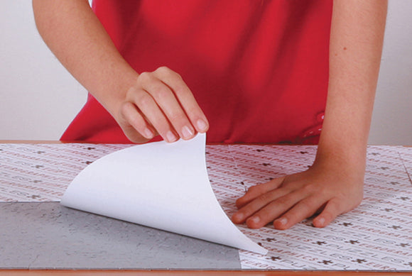 The image shows a person applying Individual Smart Puzzle Glue S, a product by Eurographics, to a jigsaw puzzle surface. The hand is seen carefully laying down a sheet of the glue on a jigsaw puzzle laid out on a surface. The puzzle features intricate designs, while the person's red shirt adds a vibrant contrast to the scene. This glue is ideal for preserving and framing completed jigsaw puzzles.