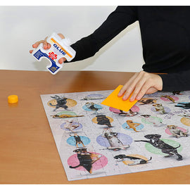 A person applying Eurographics Individual Smart Puzzle Glue on a completed jigsaw puzzle featuring various dogs. The glue bottle is in one hand, while the other hand holds a yellow smoothing tool, ensuring the pieces are secured together. The puzzle is laid out on a wooden table, showcasing colorful circular designs with different dog images.