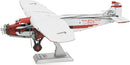 Detailed model kit of the Ford Trimotor airplane by Fascinations. The airplane features intricate design elements, including a red and white color scheme, visible engines, and landing gear. It stands on a display base, showcasing its vintage aviation charm.