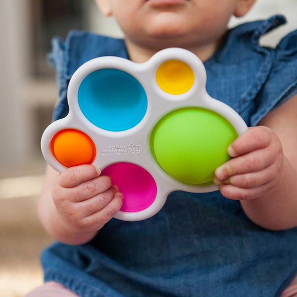 A baby holds a colorful silicone sensory toy with five different shaped buttons in vibrant colors: blue, green, orange, pink, and yellow. The toy encourages tactile exploration and provides sensory stimulation.