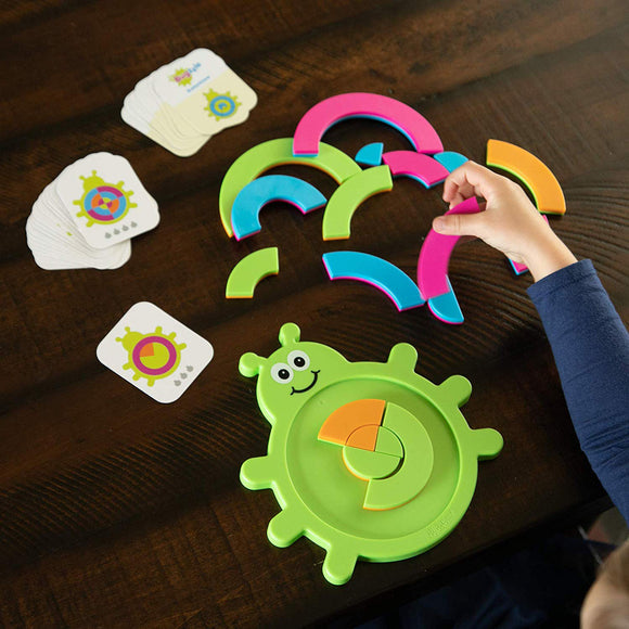 A colorful game called Bugzzle by Fat Brain Toys, featuring a green smiling bug base and various shaped pieces in bright colors including pink, blue, orange, and green. A child's hand is seen picking up a piece while several challenge cards are spread out nearby, promoting problem-solving skills and creative play.