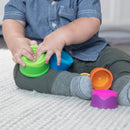 A toddler sits on a soft rug, engaging with colorful stacking cups from the Dimpl Stack by Fat Brain Toys. The child, dressed in a blue denim shirt and green pants, is focused on manipulating a green cup and a blue cup, showcasing fine motor skills. Various bright cups in orange, pink, and purple are scattered around, promoting sensory play and learning.