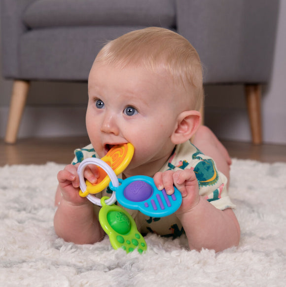 A joyful baby lying on a textured rug, holding and chewing on colorful plastic rings of the Dimpl Clutch toy by Fat Brain. The toy features various tactile elements designed for sensory exploration, with bright colors like yellow, blue, and green. The baby is focused and engaged, showcasing the playful and interactive nature of the product.