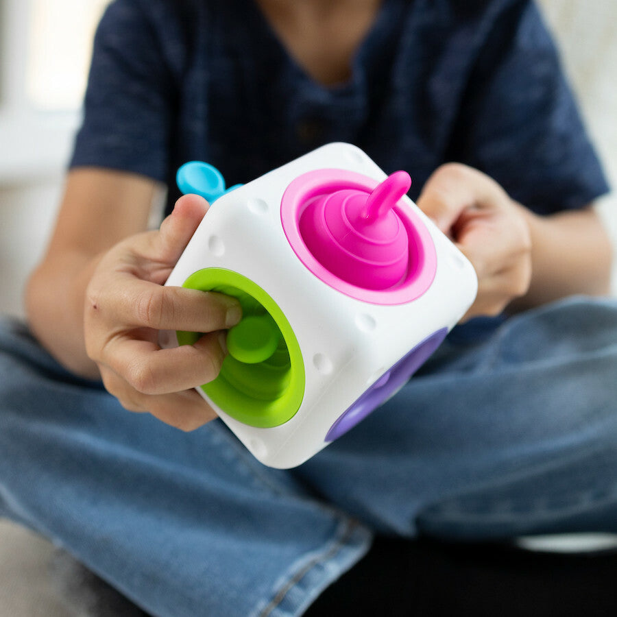 A child holds the tugl Cube made by Fat Brain, showcasing various colorful and interactive features. The cube has multiple shapes, including a pink spinner and green openings, designed for sensory play and fine motor skill development.