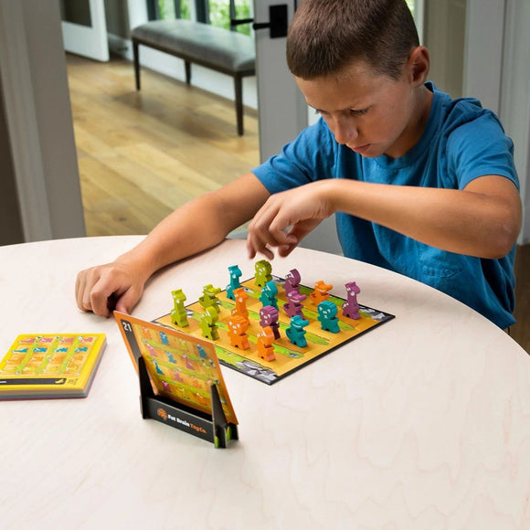 A young boy focused on playing 'Zoo Rendezvous,' a mechanical puzzle by Fat Brain. The colorful game features various animal-shaped pieces on a vibrant game board, alongside a challenge booklet. The scene is set on a light wooden table with natural lighting, highlighting the boy's concentration as he maneuvers the pieces.
