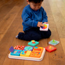 A young child seated on the floor is engaged with the Playtab Activity Board from Fat Brain. The board is colorful, featuring various interactive pieces including shapes, buttons, and sliders that promote sensory exploration and fine motor skills. The child is focused on a piece featuring bright colors, enhancing cognitive development through play.