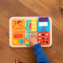 A colorful activity board from Fat Brain, featuring various tactile elements for children to explore. A child’s hand is engaging with the bright, interactive tiles designed for sensory play and fine motor skill development.