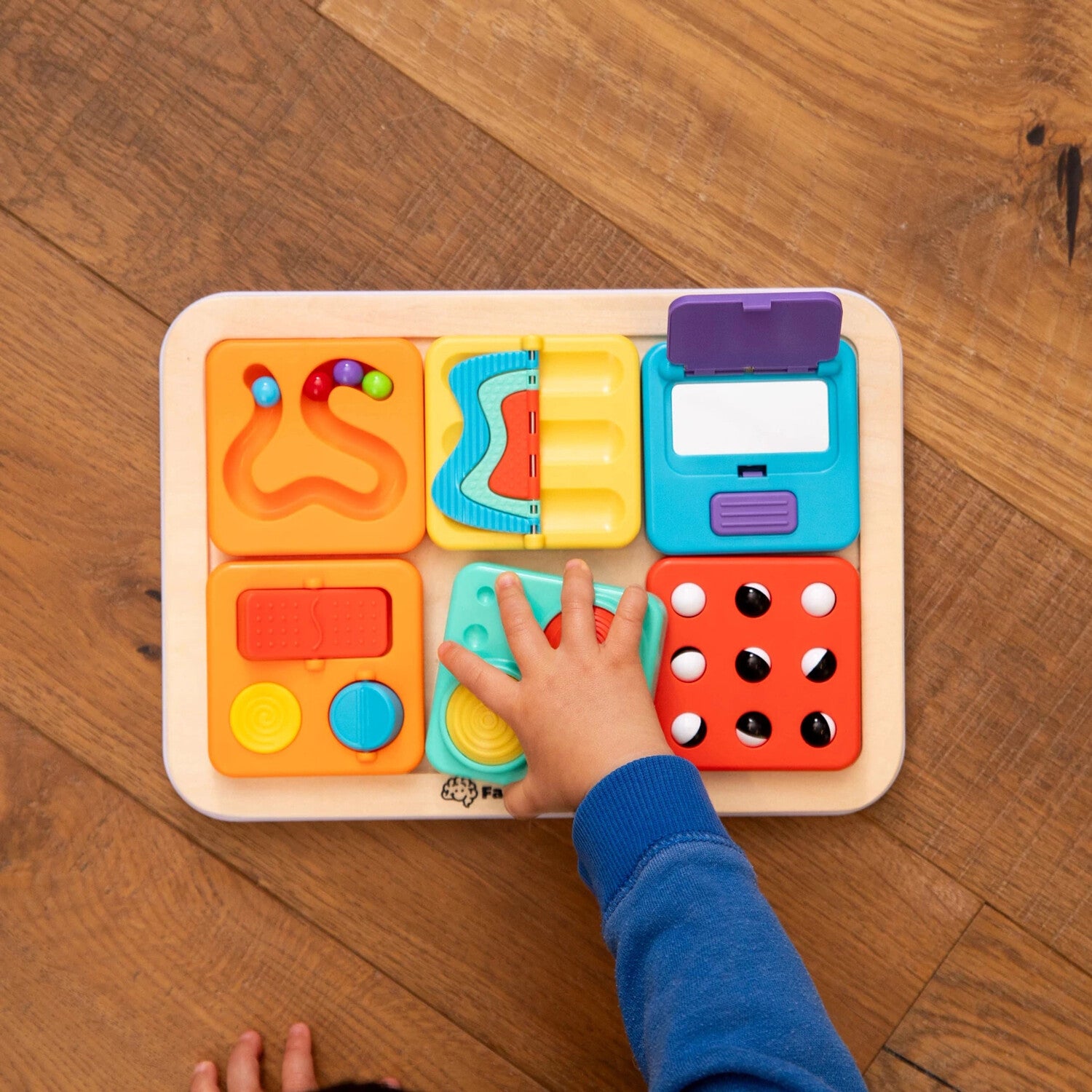 A colorful activity board from Fat Brain, featuring various tactile elements for children to explore. A child’s hand is engaging with the bright, interactive tiles designed for sensory play and fine motor skill development.