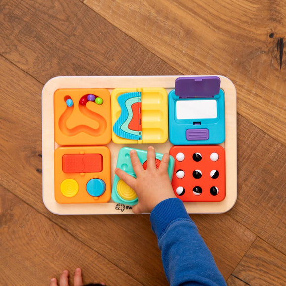 A colorful activity board from Fat Brain, featuring various tactile elements for children to explore. A child’s hand is engaging with the bright, interactive tiles designed for sensory play and fine motor skill development.