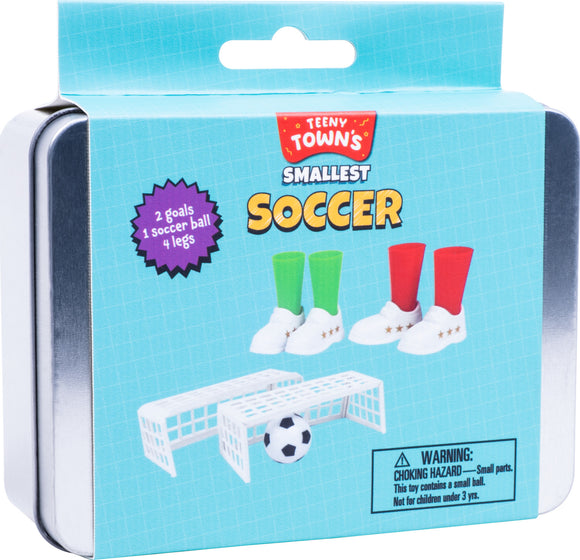 Teeny Tiny Soccer set featuring two miniature soccer goals, one small soccer ball, and four colorful player legs (two green and two red) in white shoes. The tin packaging showcases a vibrant blue background with playful graphics, warning about small parts being a choking hazard for children under 3 years old.