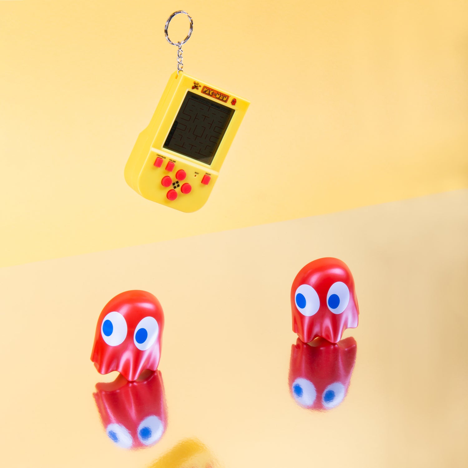 A colorful Pac-Man keyring arcade game, featuring a yellow handheld device with a small screen and buttons, hanging on a keychain. Two red ghost figures with blue eyes are placed on a reflective surface, creating a fun gaming scene.