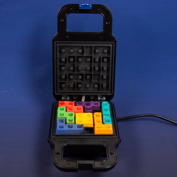 A Tetris-themed waffle maker from Fizz Creations Inc. with colorful waffle pieces resembling Tetris blocks. The device is opened, showing the waffle grid with various shapes in orange, red, purple, green, blue, and yellow, set against a blue background.