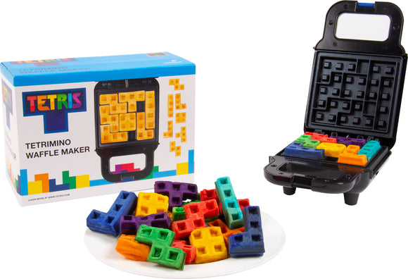 A Tetris Waffle Maker by Fizz Creations Inc. is displayed upright, with colorful Tetrimino-shaped waffles piled on a white plate beside it. The waffle maker is partially open, showcasing the vibrant waffles inside and the branded box in the background, featuring the iconic Tetris logo and colorful design.