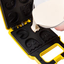 A person pours pancake batter into a yellow Pac Man waffle maker featuring iconic Pac Man and ghost shapes on the cooking plates. The waffle maker is open, revealing multiple molds for creating themed waffles.