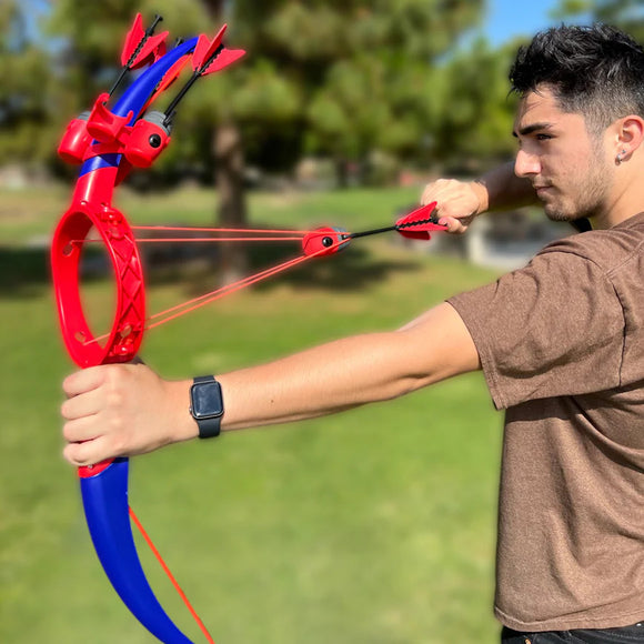 A young man stands outside holding the Ring Strike game by Funwares, which features a vibrant red and blue color scheme. He is aiming the uniquely designed toy slingshot, which resembles a bow with four pointed projectiles ready to be launched. The background shows a grassy field, indicating an outdoor play setting, perfect for social games and friendly competition.