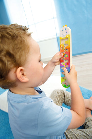 A young child with curly hair sits on a blue mat, engrossed in playing with the Rainmaker Wormy toy from Haba. The colorful toy features a smiling face and beads that move through a winding, transparent path as the child tilts it. The child holds the toy securely, showcasing its engaging design and educational features that promote fine motor skills and sensory play.