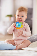 A happy baby sitting on a soft surface, holding the colorful Haba Grasping Toy Blossom. The toy features a round center and multiple colorful textured segments, designed to stimulate sensory play. The baby, wearing a light pink onesie with polka dots, is smiling and playfully engaging with the toy.