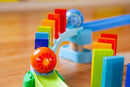 The Kubu Ball Track Kringel Domino from HABA features colorful wooden blocks and a vibrant, rolling ball. The setup showcases a playful path where the ball can roll through various routes, knocking down rainbow-colored dominos, facilitating hands-on creative play and developing fine motor skills for children.