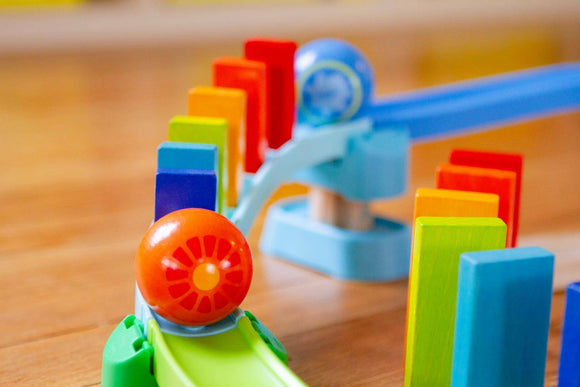 The Kubu Ball Track Kringel Domino from HABA features colorful wooden blocks and a vibrant, rolling ball. The setup showcases a playful path where the ball can roll through various routes, knocking down rainbow-colored dominos, facilitating hands-on creative play and developing fine motor skills for children.