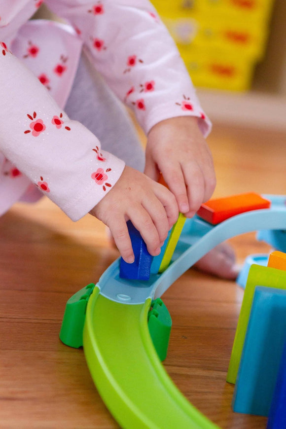 A child’s hands placing colorful blocks onto a Kubu Ball Track Kringel Domino kit made by HABA. The vibrant components are set on a wooden floor, showcasing the interactive features of the DIY toy.