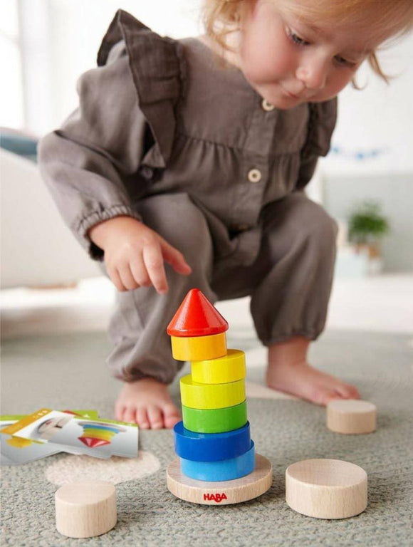 A child dressed in gray is skillfully stacking colorful wooden rings in the Stacking Game Wobbly Tower by Haba. The game features a vibrant tower of rings in various colors including red, yellow, green, and blue, set on a circular wooden base. Nearby, a couple of circular wooden pieces are visible, adding to the play setup.