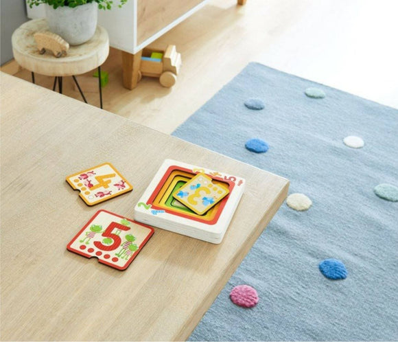 A colorful wooden puzzle titled 'Counting Friends' by HABA. The puzzle pieces, featuring numbers 4 and 5 with playful designs of animals, are displayed on a light wooden table. The setting includes a cozy rug with colorful dots, enhancing the playful atmosphere. Ideal for developing counting skills and enhancing fine motor abilities in young children.