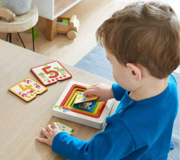 A child wearing a blue shirt is engaged with the HABA Wooden Puzzle Counting Friends on a wooden table. The puzzle features colorful pieces with numbers 4 and 5 prominently displayed, while the child is inserting a puzzle piece. The background shows a bright, inviting playroom setting.