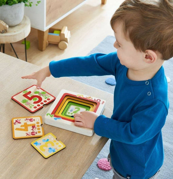 A young child engaging with the Wooden Puzzle Counting Friends by HABA. The child is placing number pieces into a wooden base that features colorful illustrations. The puzzle includes numbers from 1 to 5 with various designs on each piece. The setting is a light and playful room with colorful decor.